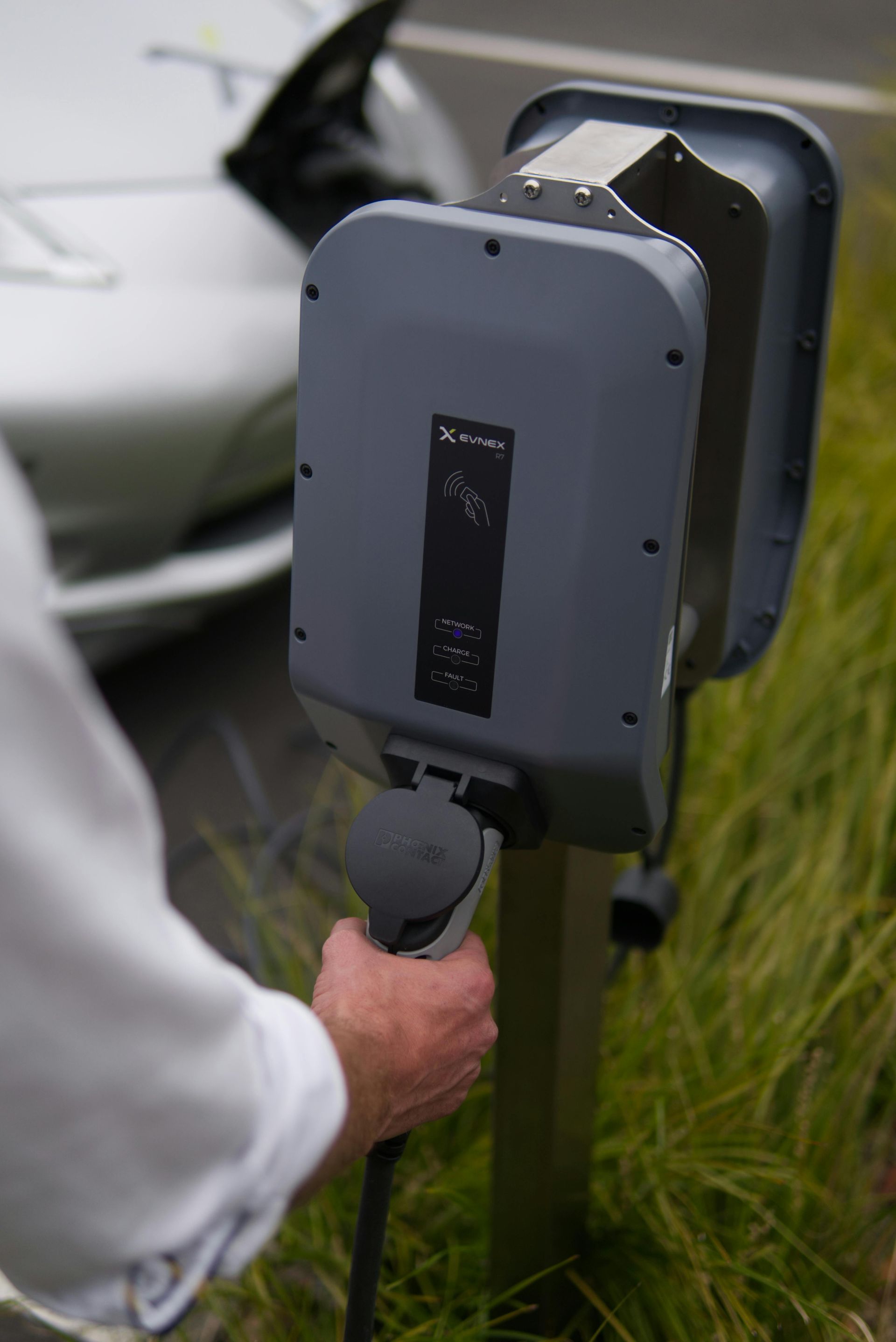 Person plugging an electric vehicle charger into a gray charging station next to a car.