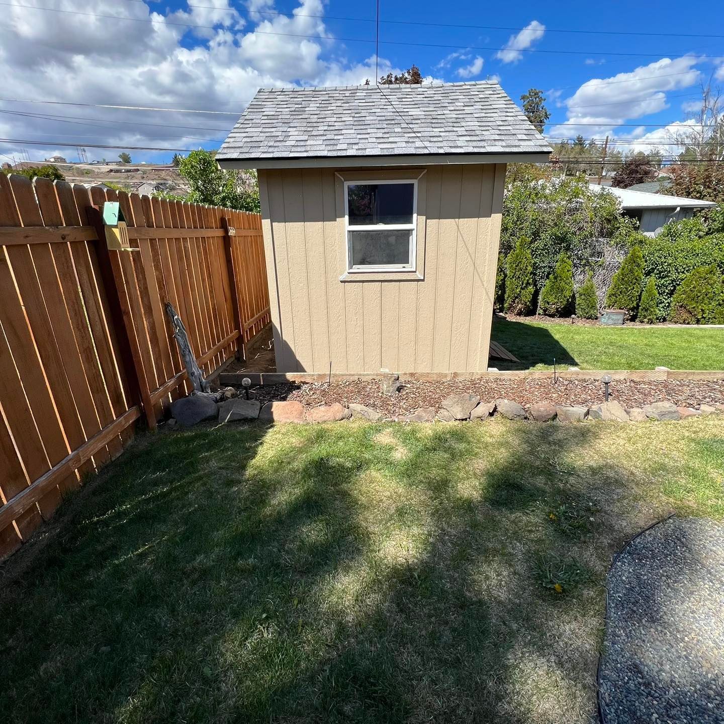 A small shed with a window is in the backyard next to a wooden fence.