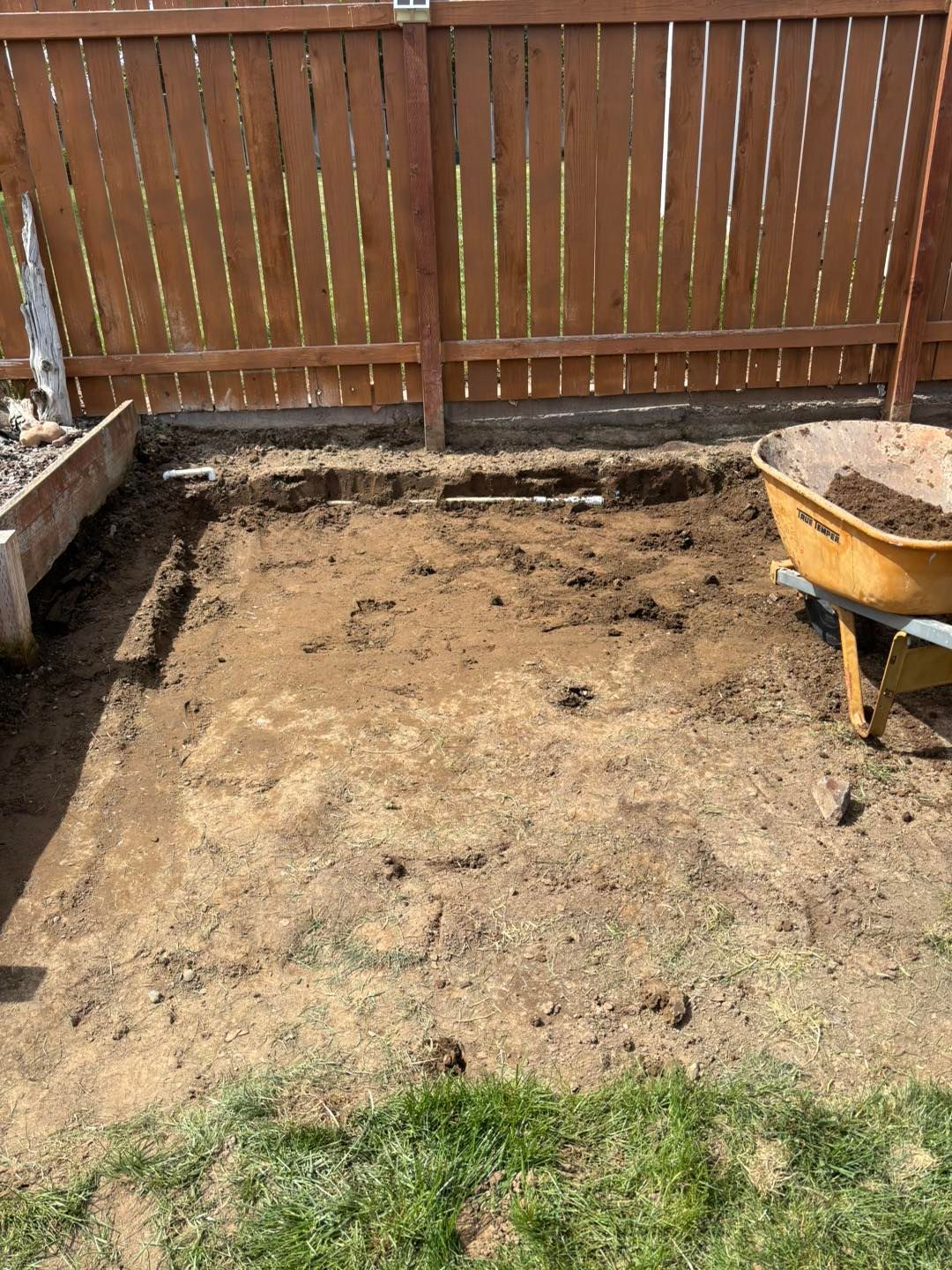 A wheelbarrow filled with dirt is sitting in a yard next to a wooden fence.