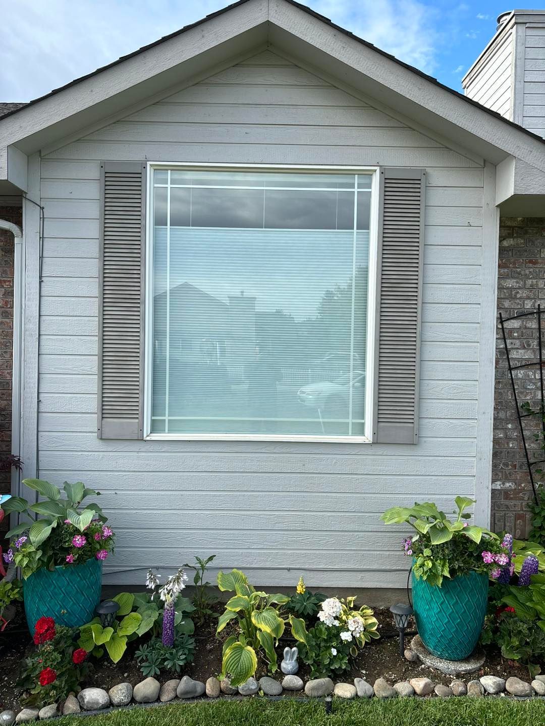 A white house with a large window and shutters surrounded by potted plants.