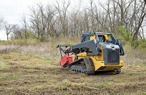 A yellow and gray compact track loader with a forestry mulcher attachment working in a field with trees in the background.
