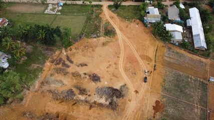 An aerial view of a dirt clearing in a rural setting, with tire tracks leading toward nearby homes and green fields.