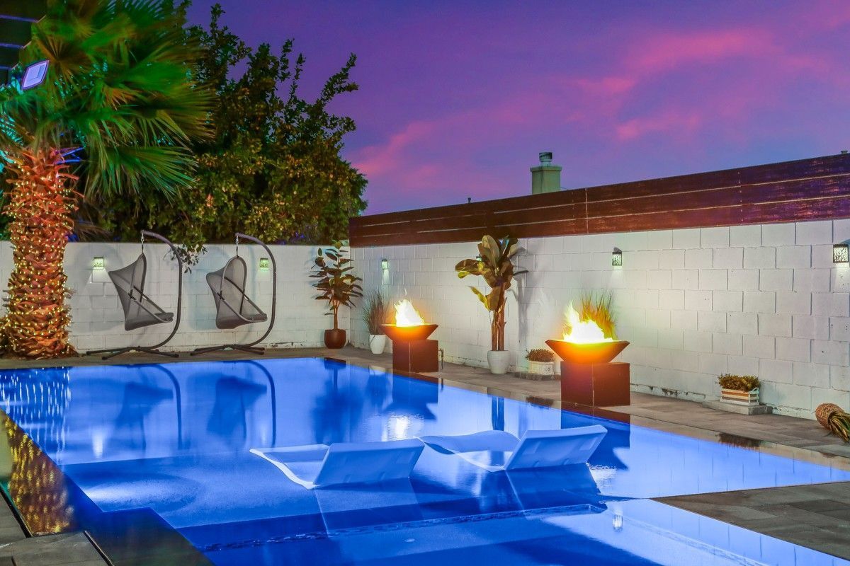 Poolside nighttime scene with a pool, fire pits, and hanging chairs under a colorful sky.