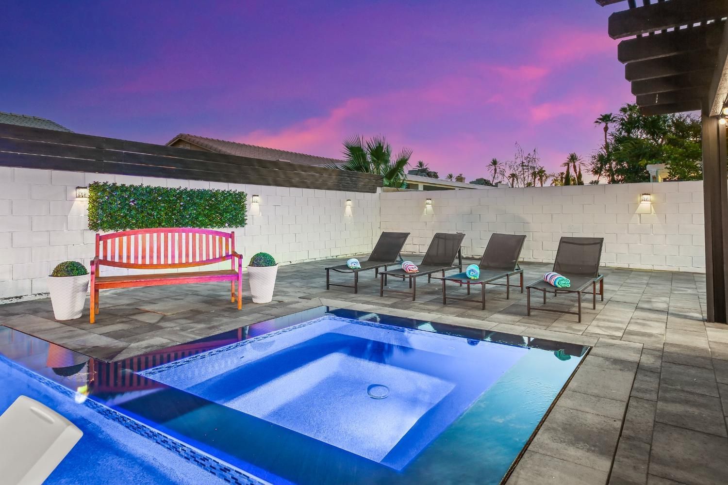 Pool and spa at dusk; lounge chairs, bench, and wall with green hedge under a colorful sky.