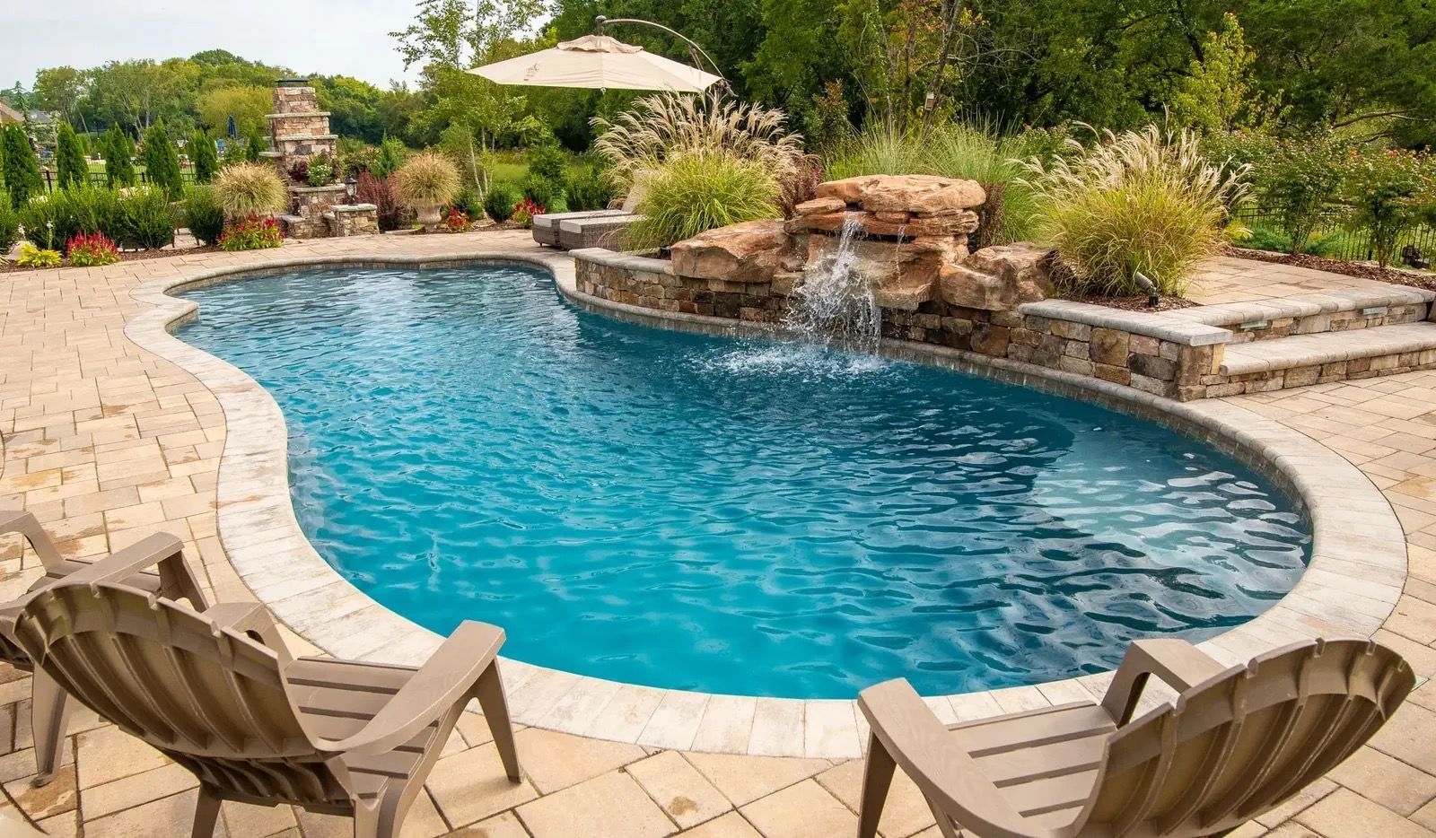 A backyard swimming pool with waterfall feature, surrounded by stone patio and lounge chairs.