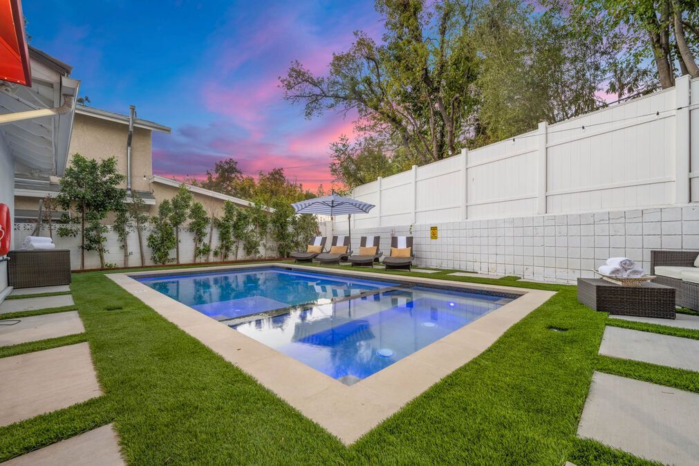 Backyard pool with lounge chairs under an umbrella, lush green grass, and a colorful sunset.