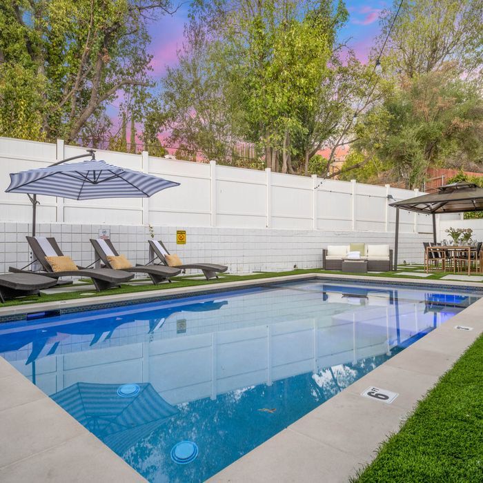 Swimming pool with lounge chairs, umbrella, and dining area under a gazebo; white fence, blue sky.