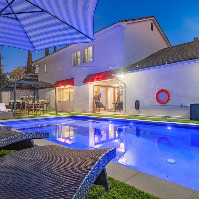 Backyard pool at night with two-story house, outdoor dining, and lounge chairs. Blue pool lights.