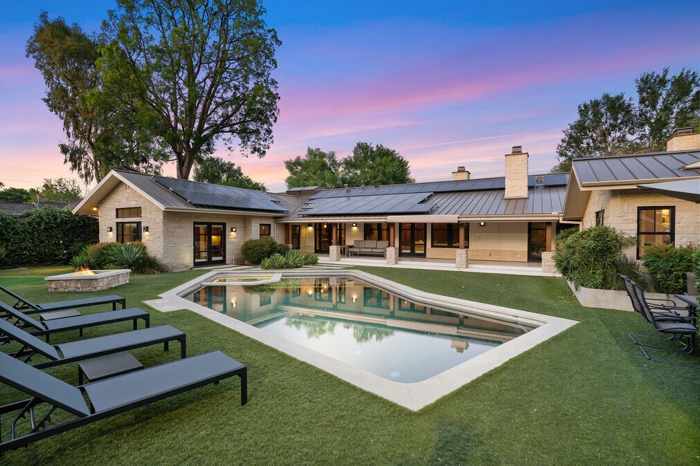 Backyard with pool, lounge chairs, and house with stone facade at dusk under a colorful sky.