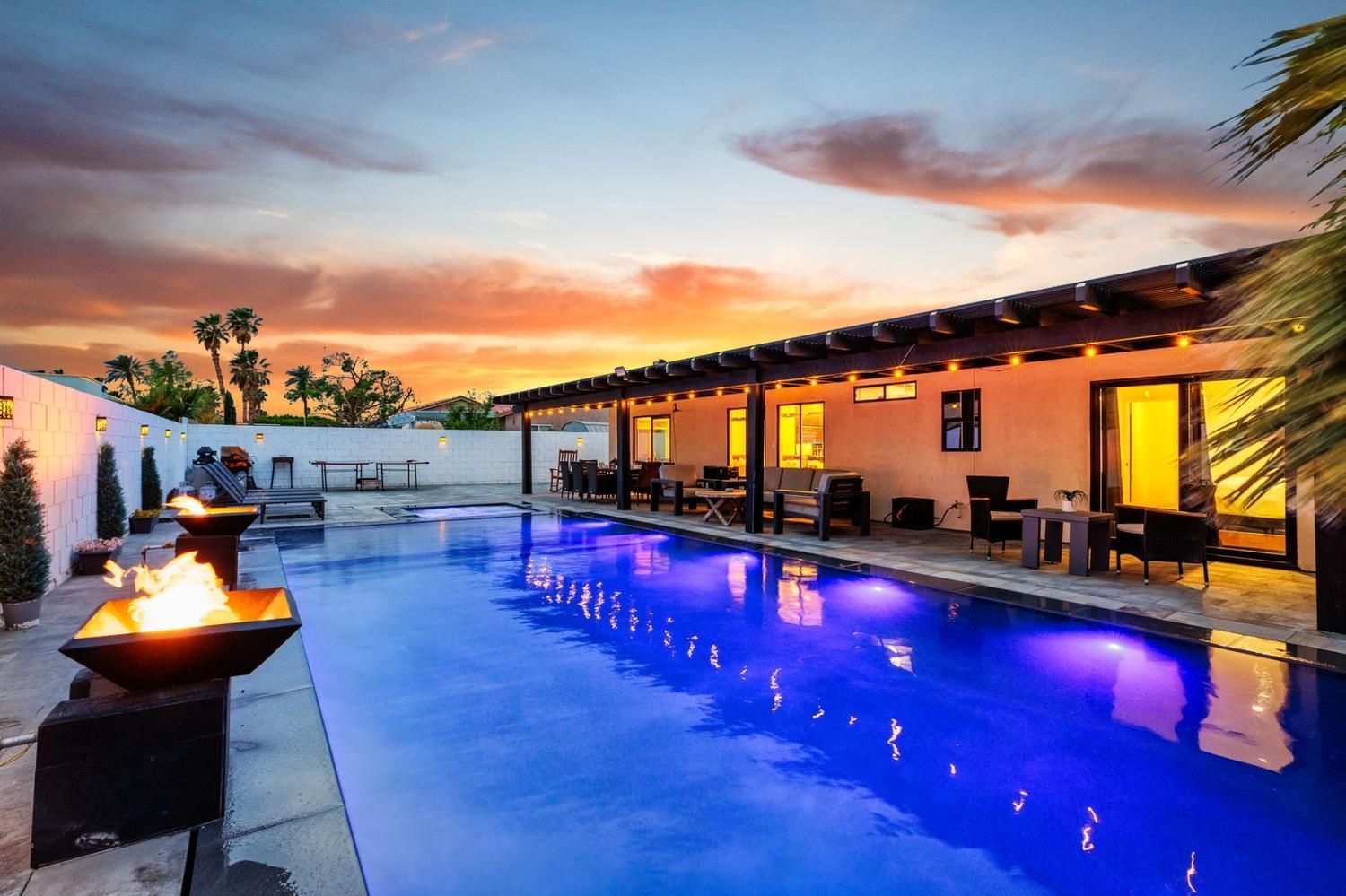 Poolside evening scene: long pool with blue water, fire pit, patio, sunset.
