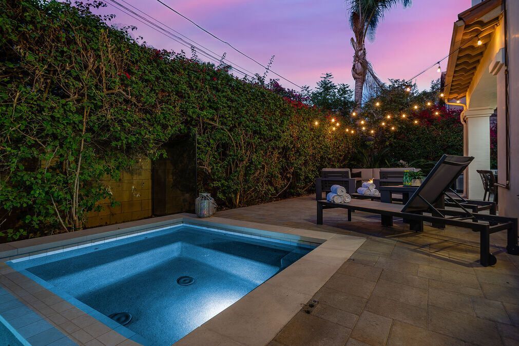 Hot tub and patio at dusk; string lights, lush greenery, and lounge chairs.
