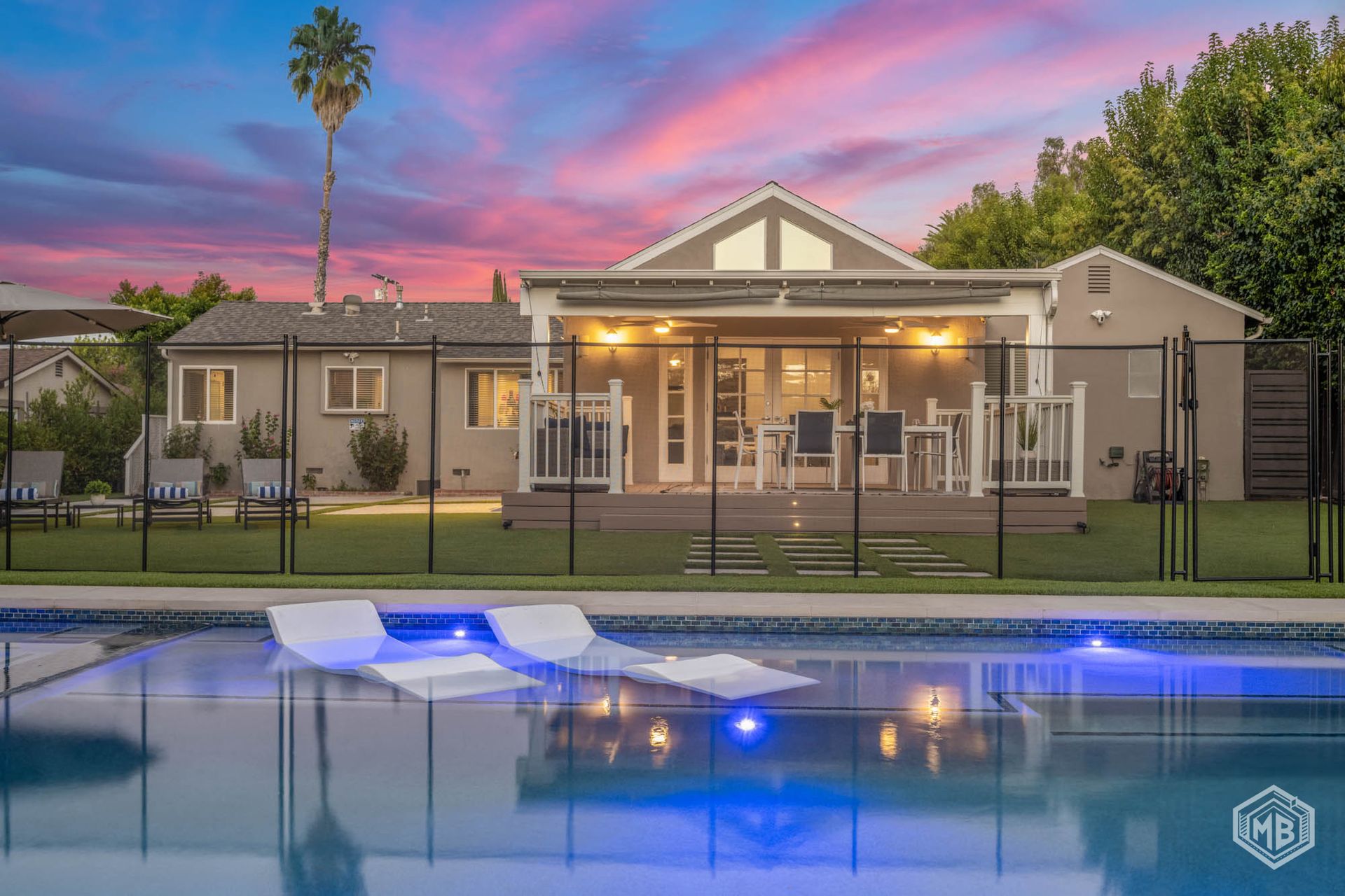 Pool and spa at dusk; lounge chairs, bench, and wall with green hedge under a colorful sky.