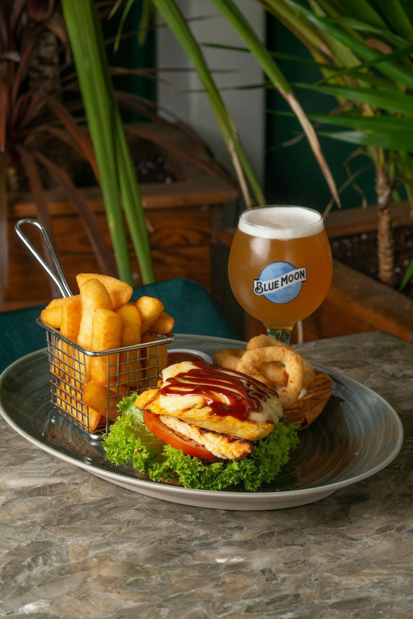 A plate of chicken burger, onion rings, and a basket of fries served with a glass of Blue Moon beer on a stone table.