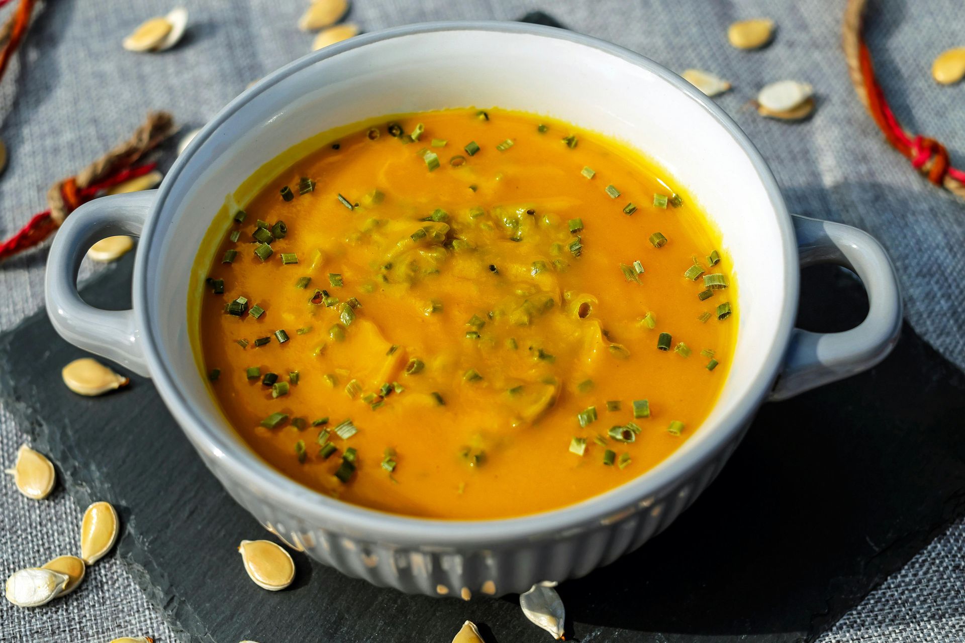 A bowl of orange pumpkin soup garnished with green herbs, set on a dark slate mat surrounded by scattered pumpkin seeds.