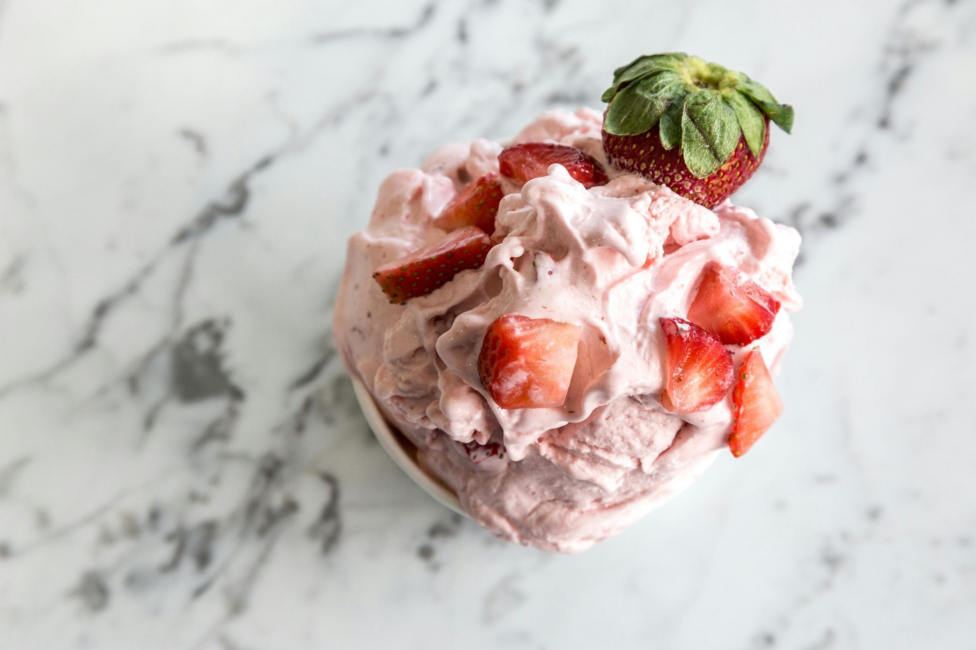 A bowl of creamy strawberry ice cream topped with fresh strawberry slices, set on a white marble surface.