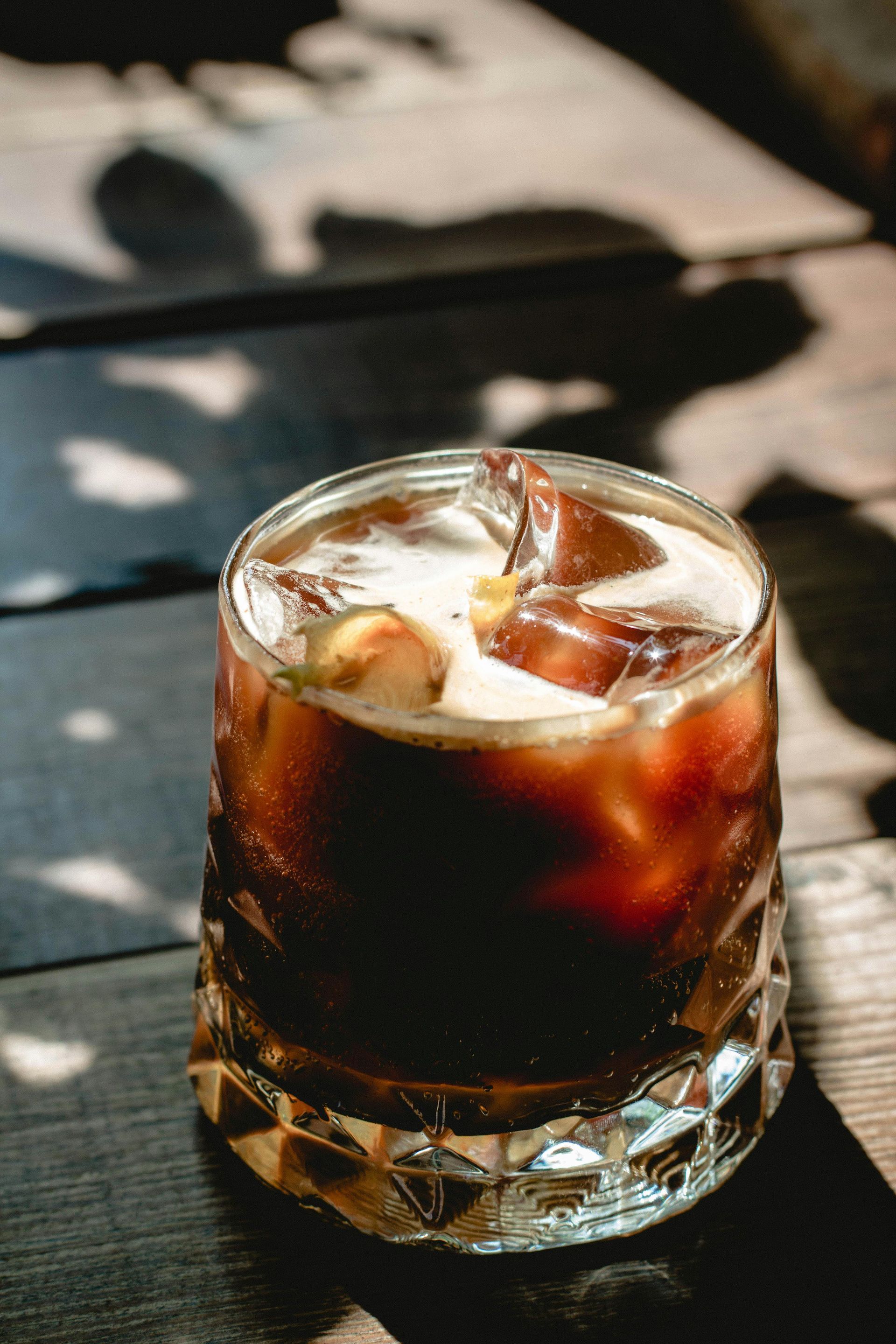 A glass of iced coffee in a decorative crystal tumbler on a wooden surface, with light casting leaf shadows nearby.