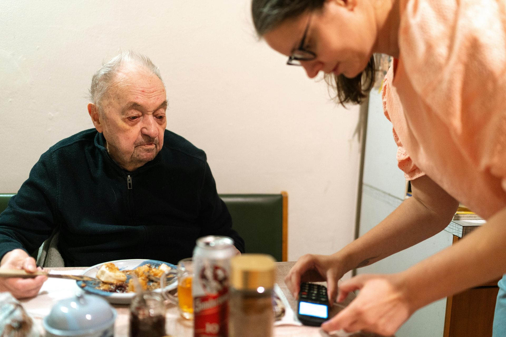 A person standing at a table uses a small electronic device while another person sits and eats a meal.