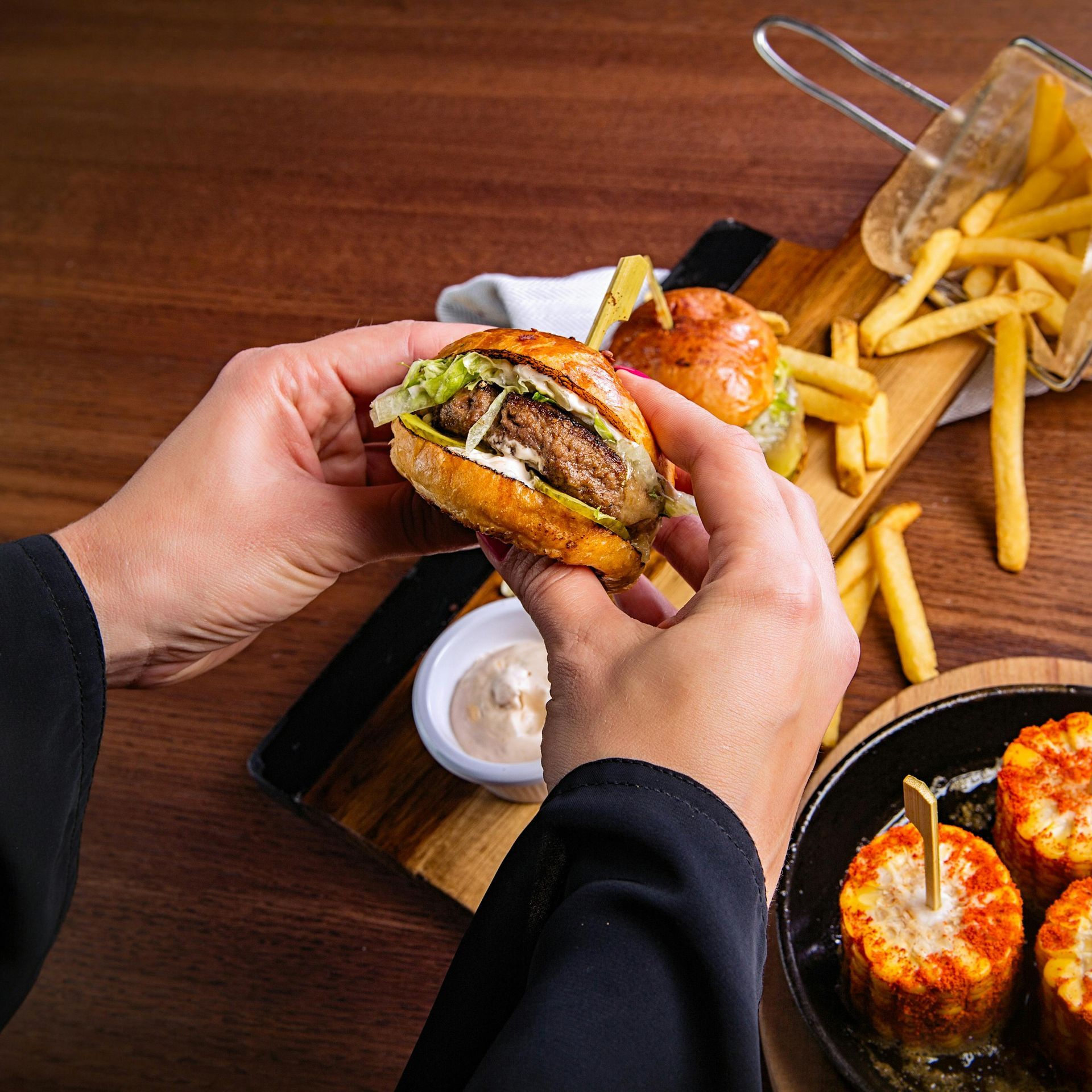 Hands hold a mini burger above a wooden serving board with fries, a dipping sauce, and corn-on-the-cob skewers.