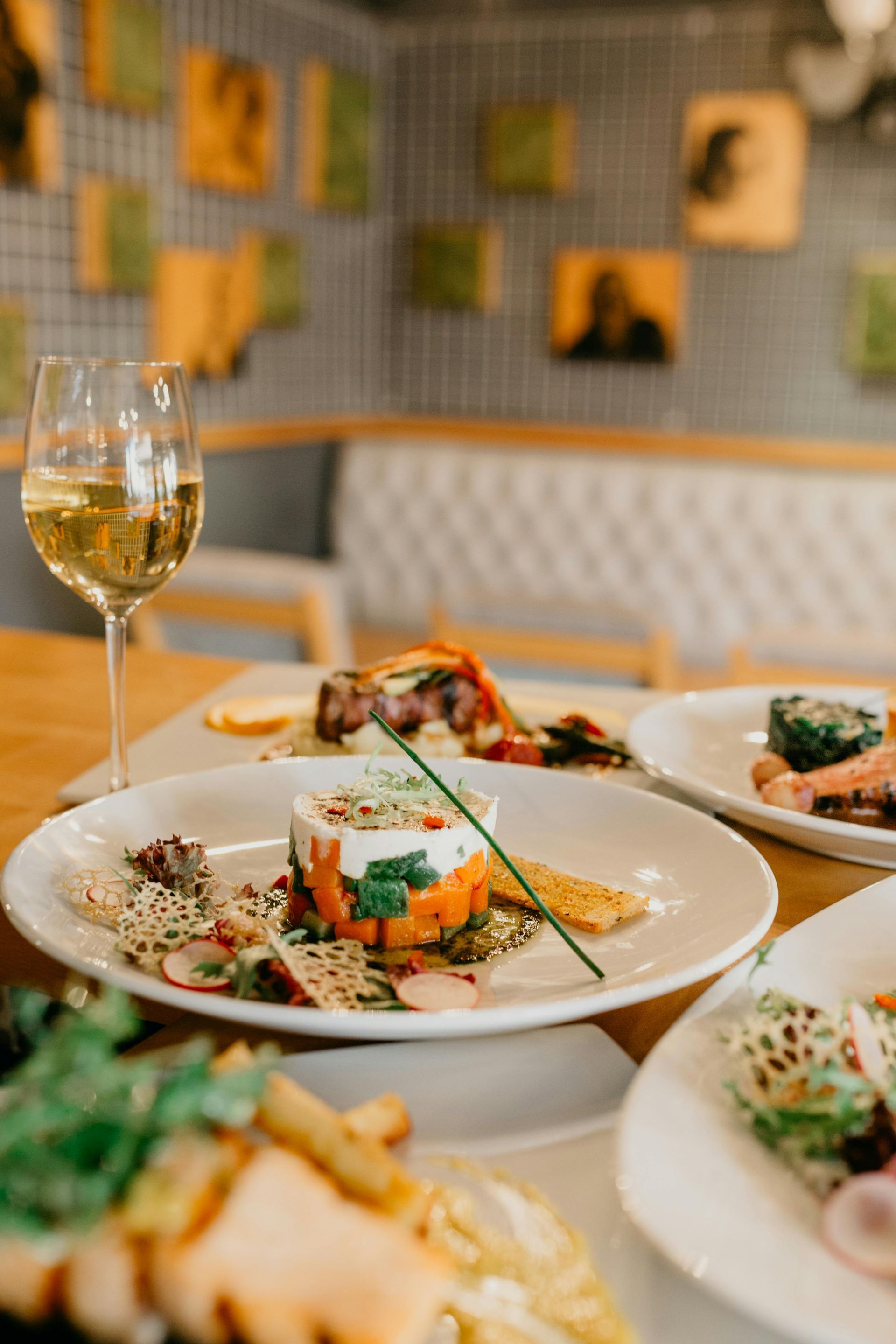 Plated gourmet appetizers, a glass of white wine, and blurred seating in a dimly lit, contemporary restaurant setting.