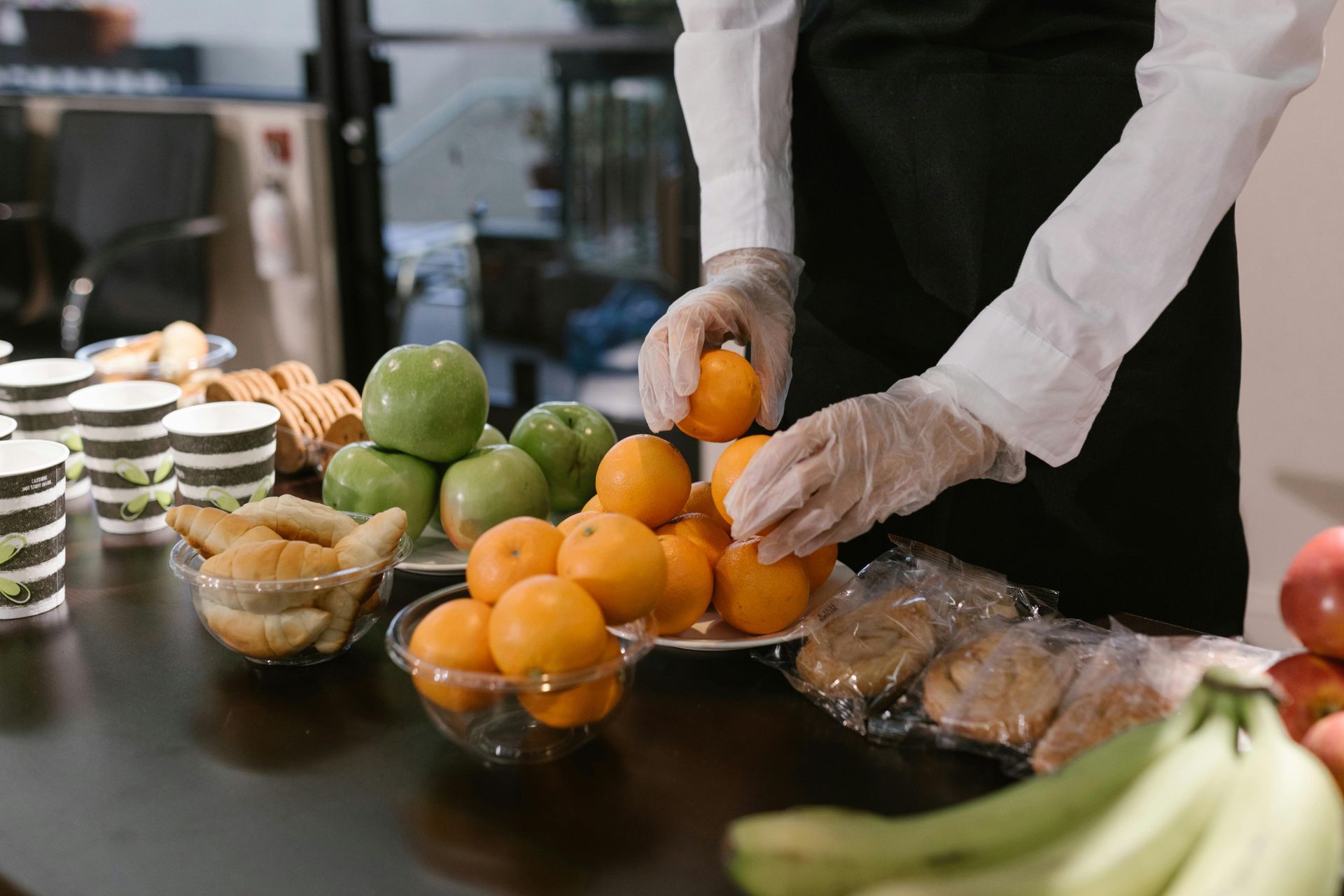 A server wearing gloves arranges fresh oranges on a table alongside bowls of croissants, apples, and bananas.