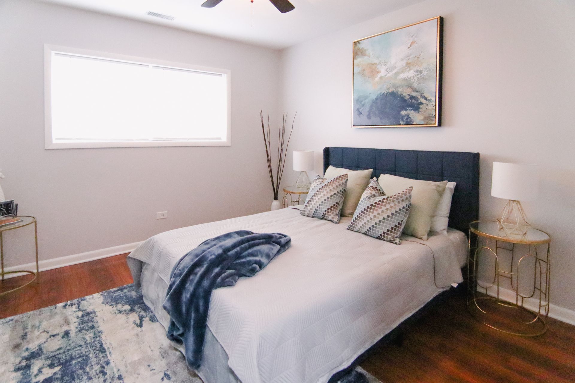 Bedroom with bed, art, rug, bedside tables; neutral tones, blue accents.