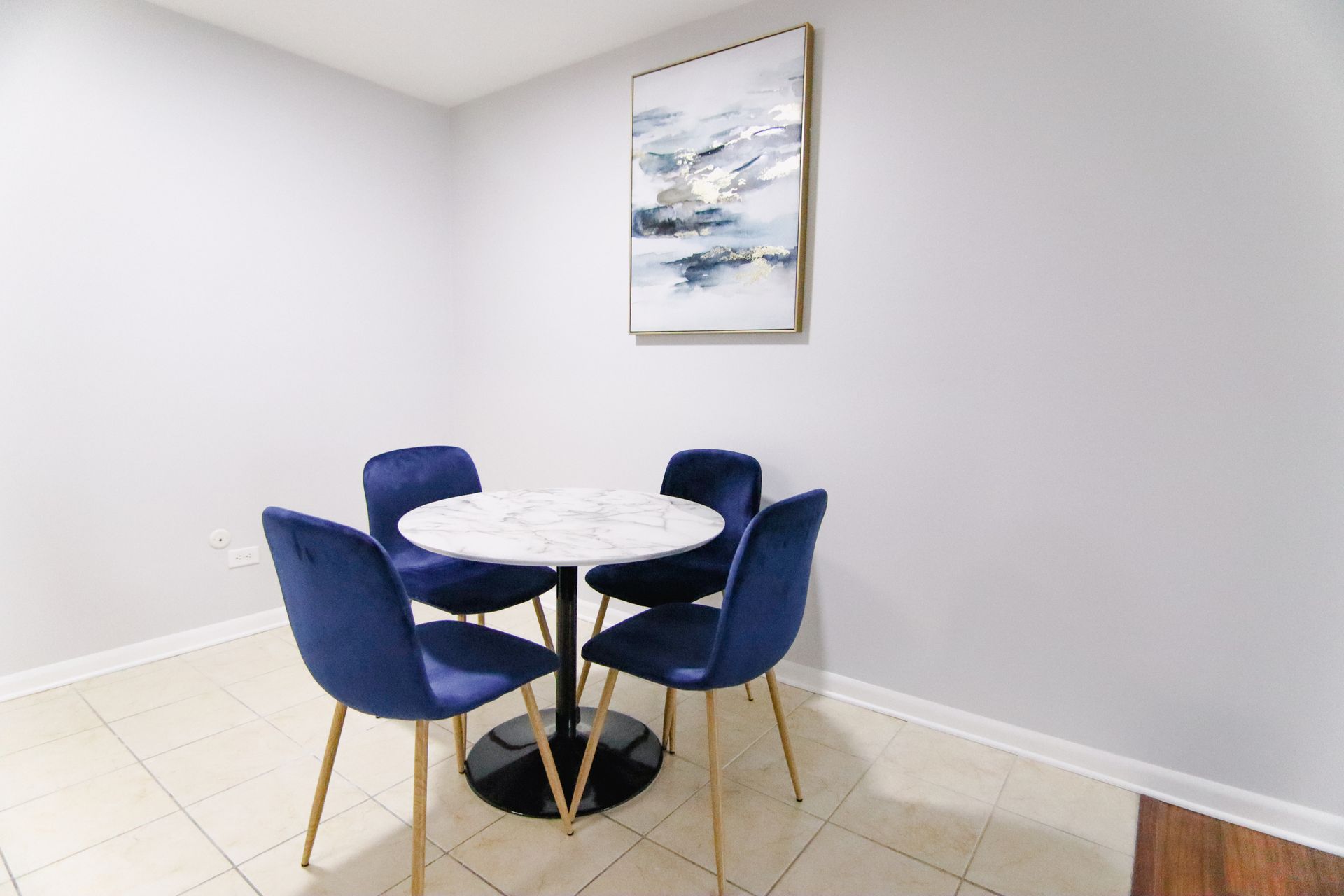 A small dining area with a round marble table, four blue velvet chairs, and abstract wall art.