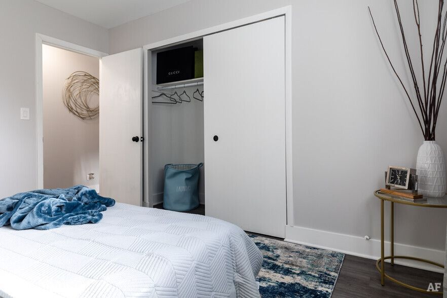 Bedroom with white closet doors, bed, and vase with sticks. Gray walls, rug, and small table.