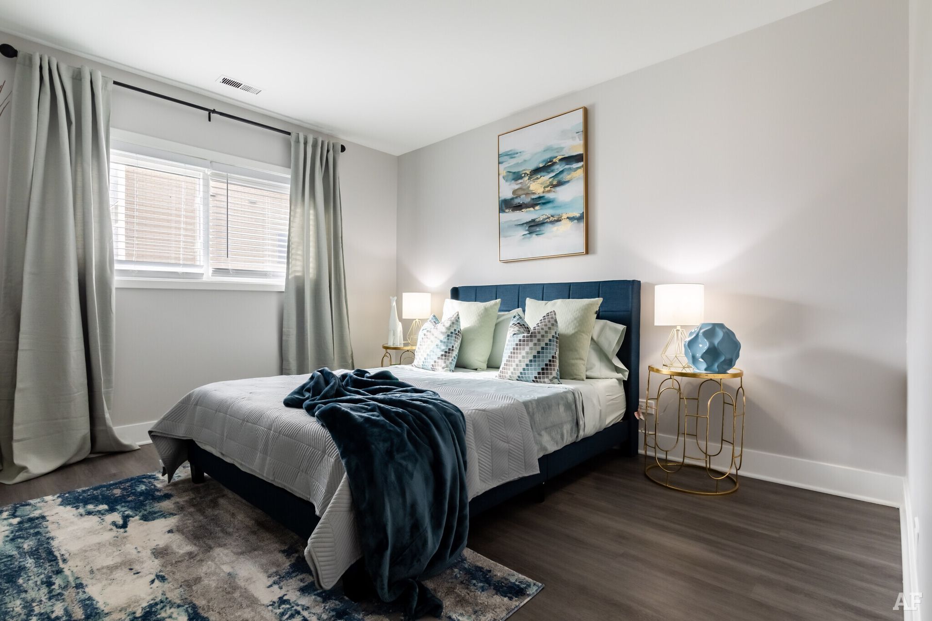 Bedroom with blue bed, curtains, and rug; artwork on the wall, a window, and two bedside tables.