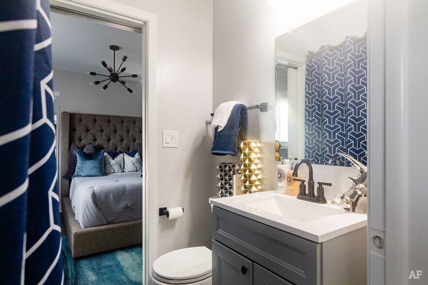 Bathroom and bedroom interior. Gray vanity and wall, blue and white patterned shower curtain. Bedroom has blue bed.