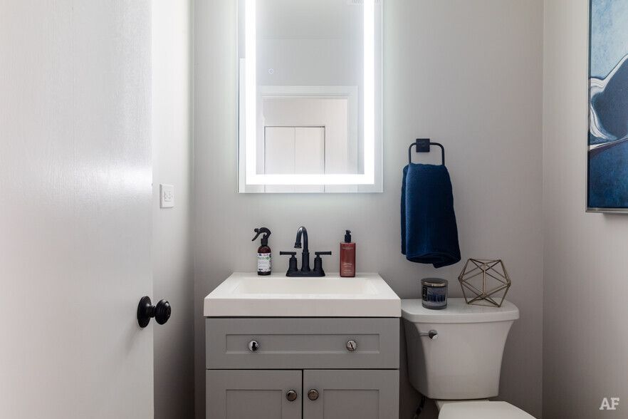 Small gray bathroom with vanity, toilet, and lighted mirror; navy blue towel.