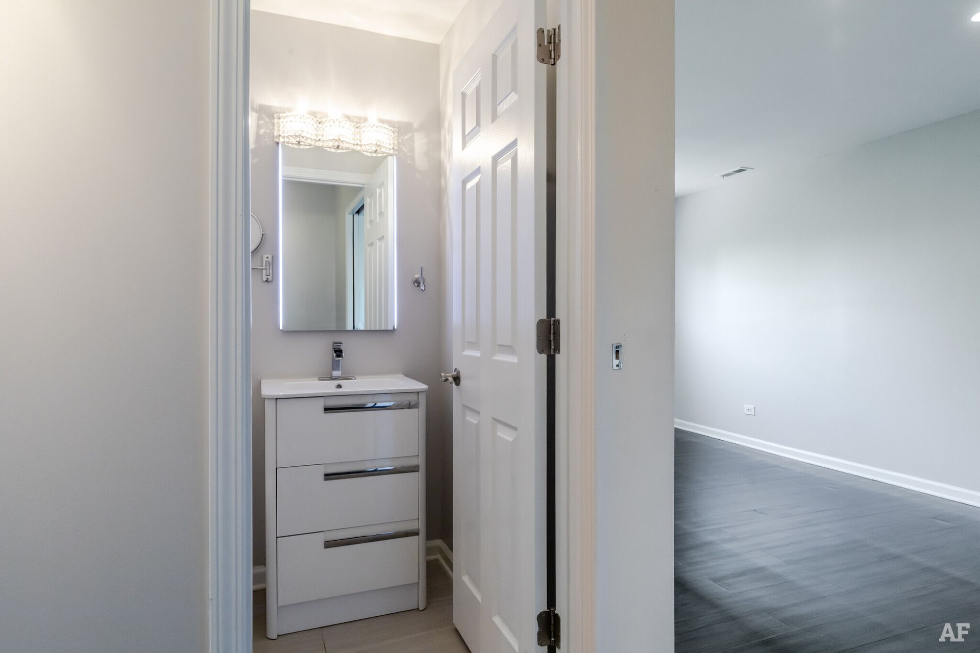 Bathroom with white vanity, mirror, and doorway to a room with dark flooring.
