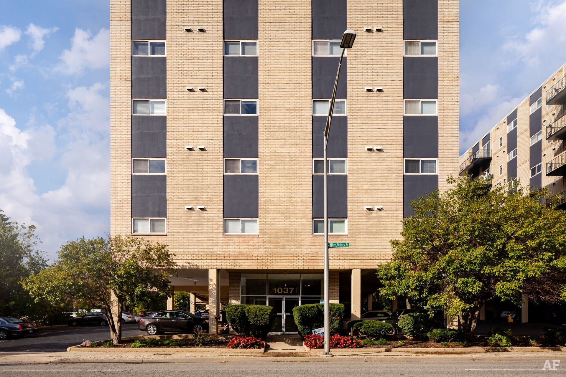 A tall apartment building with dark vertical panels and light brick exterior, green bushes in front.