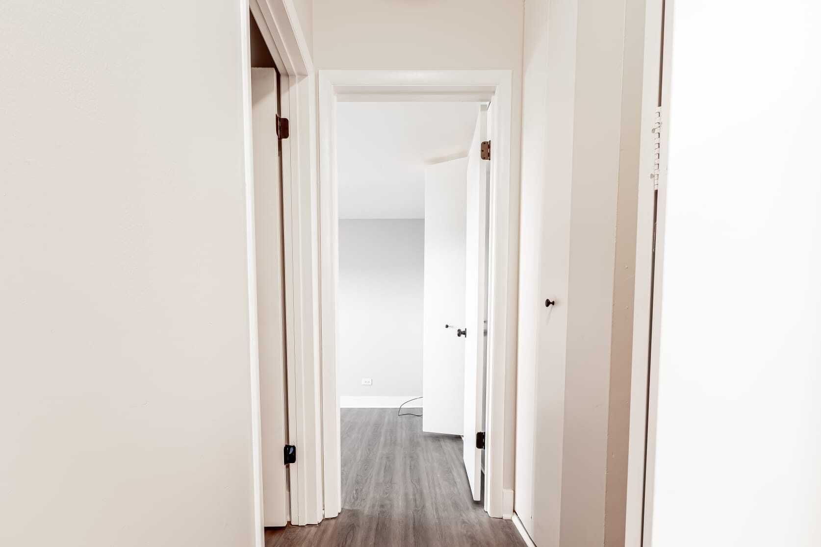 Bright hallway with white walls, doors, and gray flooring. The hallway leads to a room with a closed door.