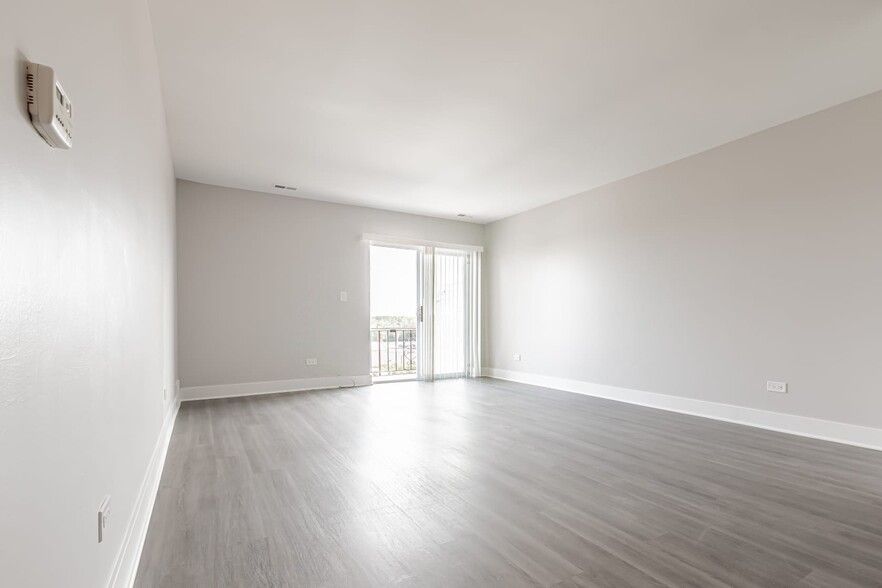 Empty, light-filled room with gray wood-look floor. Sliding glass doors lead to a balcony. White walls, neutral tones.