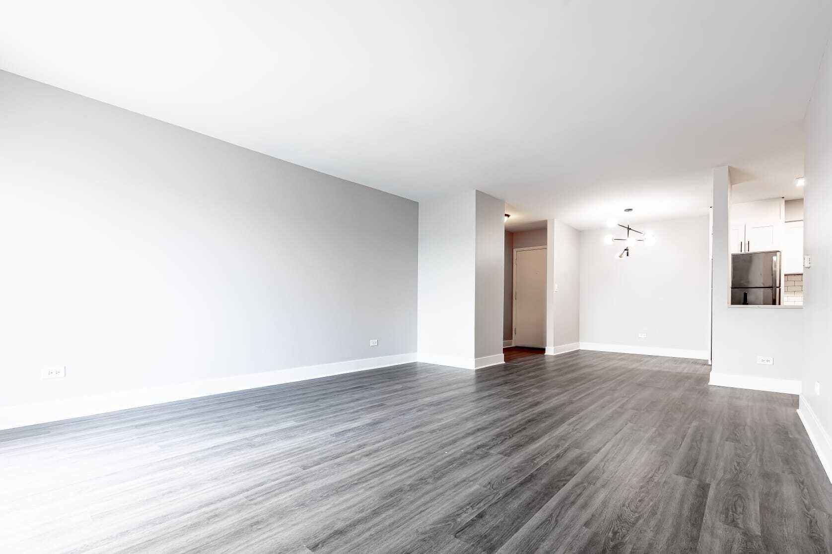 Empty living room with gray walls, dark wood floors, and a doorway to a kitchen.