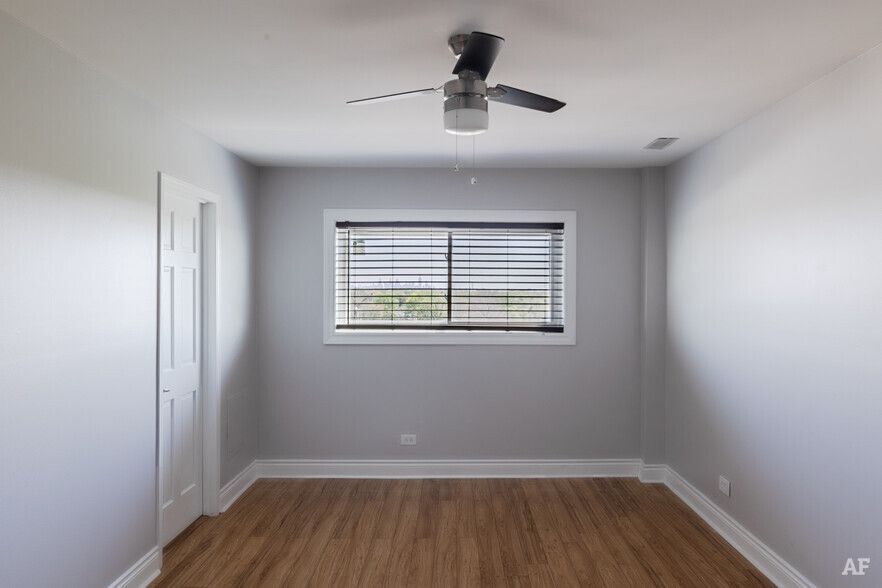 Empty room with gray walls, wood floors, and a window with blinds. A ceiling fan hangs overhead.