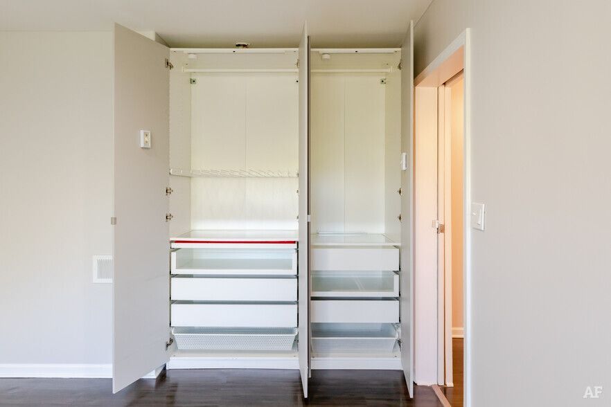 Open white closet with drawers and hanging rods in a room with wood floors and a doorway.