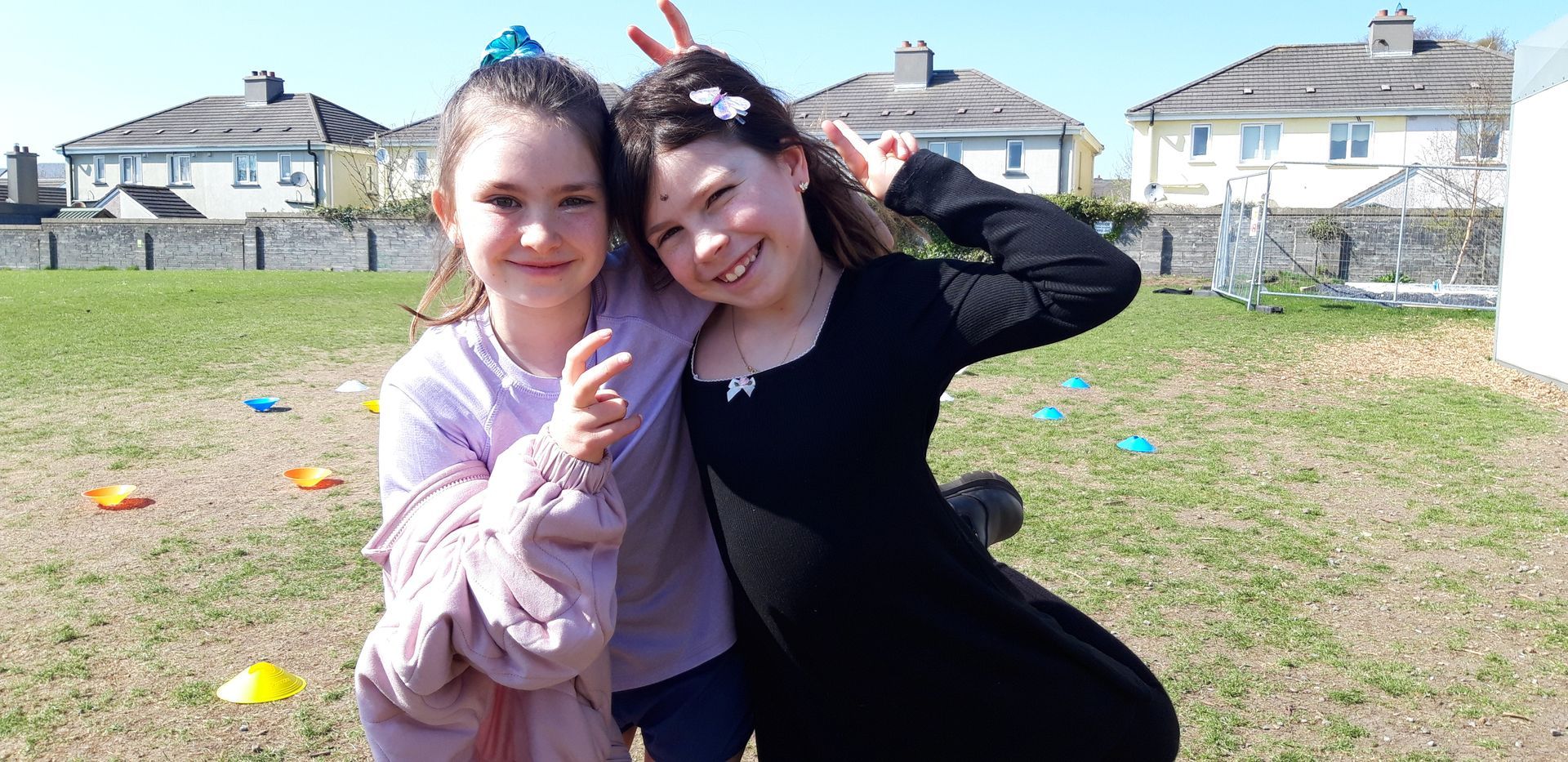Two young girls posing for a photo outdoors. One points with a smile, the other makes a peace sign.