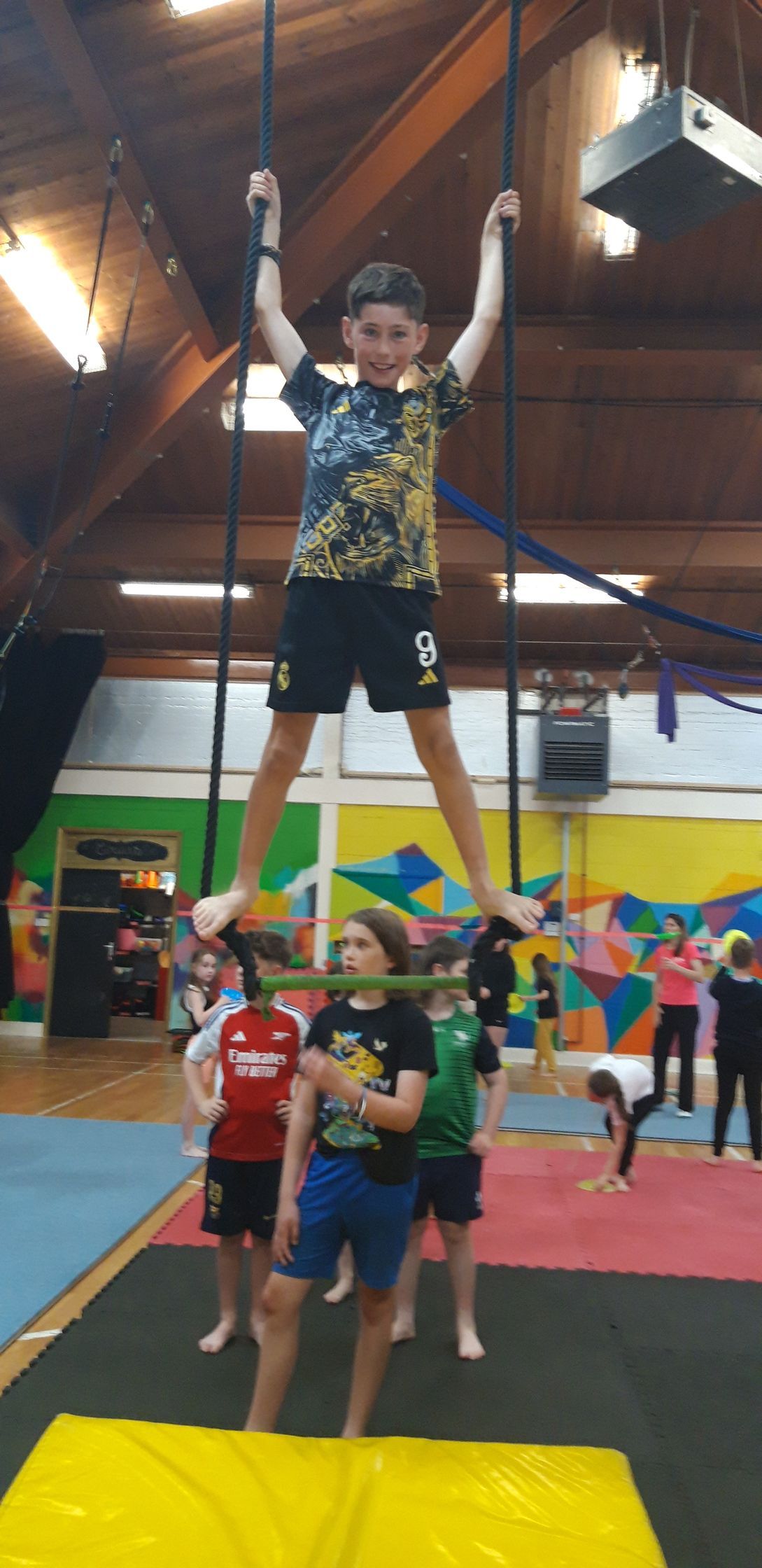 Boy balances on horizontal bar held by other children, hanging from ropes indoors.