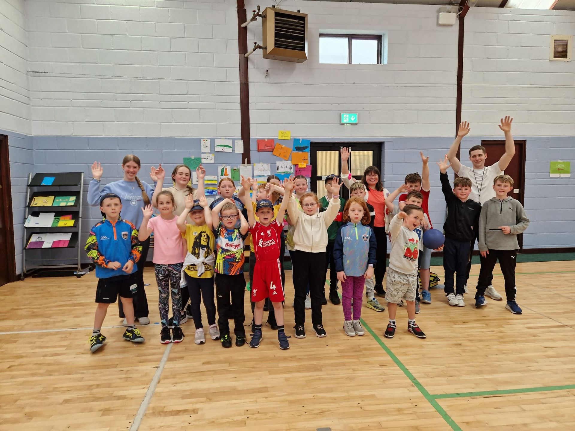 Group of children and adults in a gymnasium waving, smiling. Wooden floor, colorful posters, and shelving.