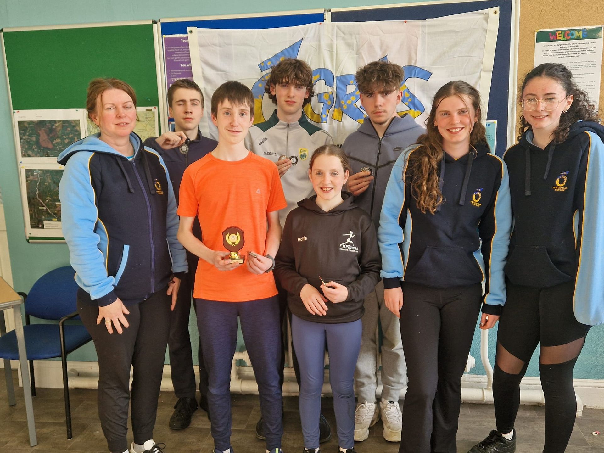 Grúpa daltaí agus múinteoir ag seasamh laistigh, duine acu ag coinneáil trófaí beag os comhair cláir fógraí.