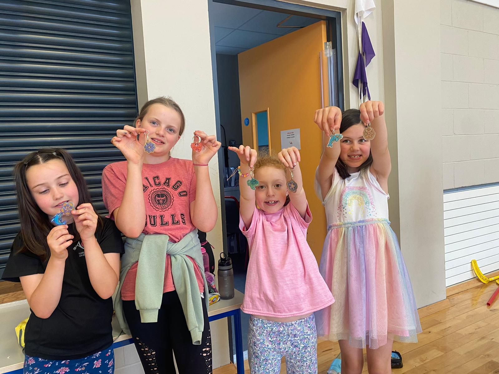 Four smiling girls hold up crafts inside, showing off their creations.