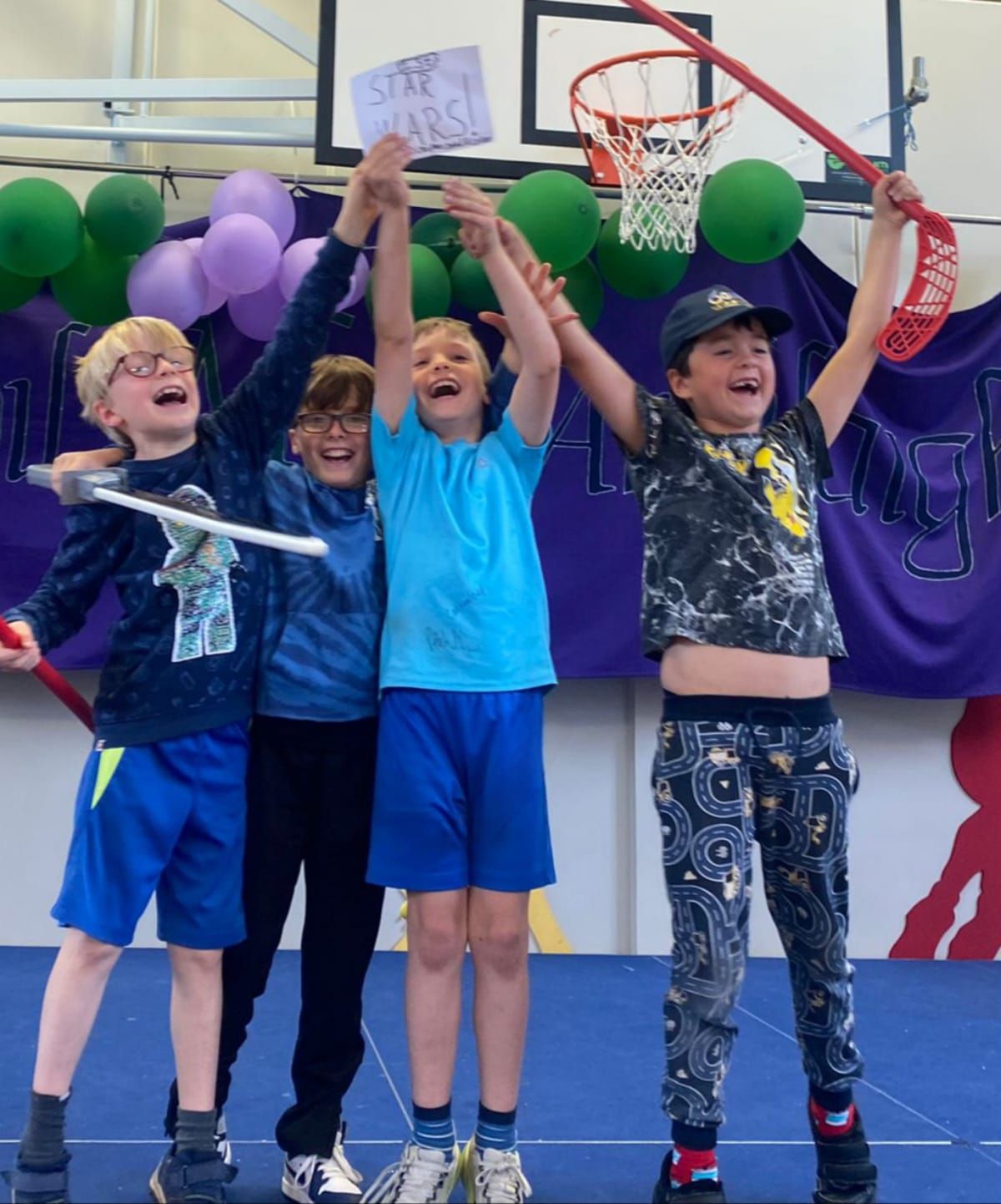 Four kids cheering, holding up sticks and sign saying “STAR WARS!” in a gym with balloons and a basketball hoop.
