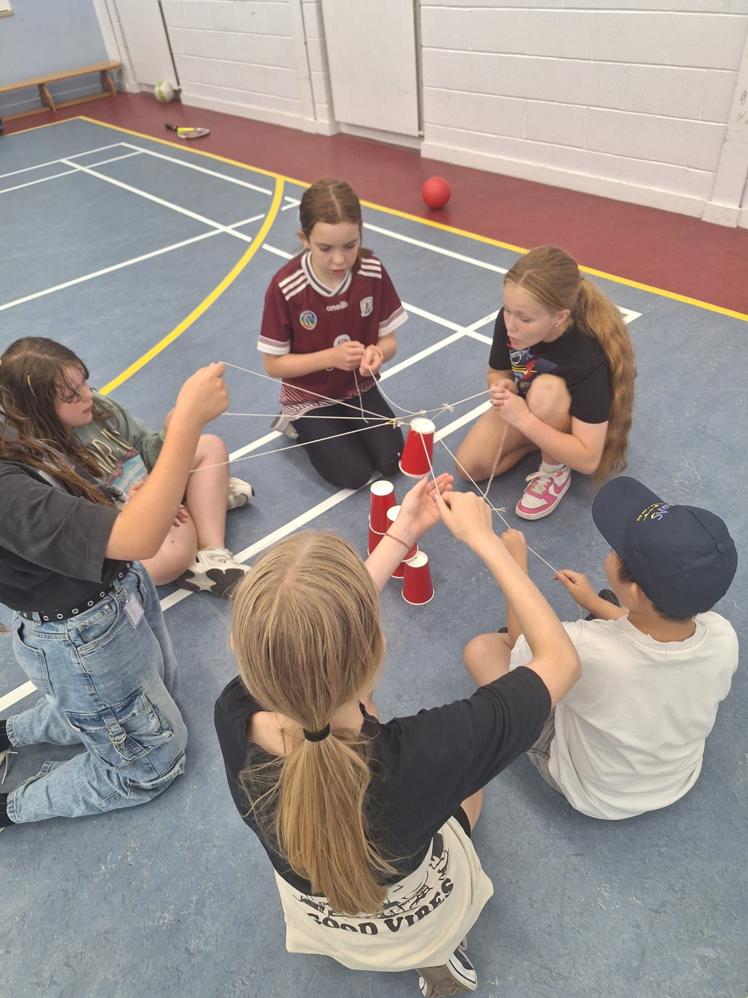 Six children seated in a circle, using strings to manipulate red cups, indoors on a blue sports court.