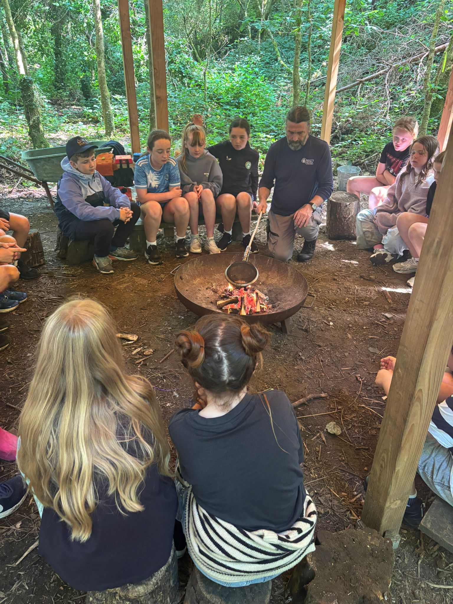 Group of people around a fire pit in a wooded area, with a person demonstrating.