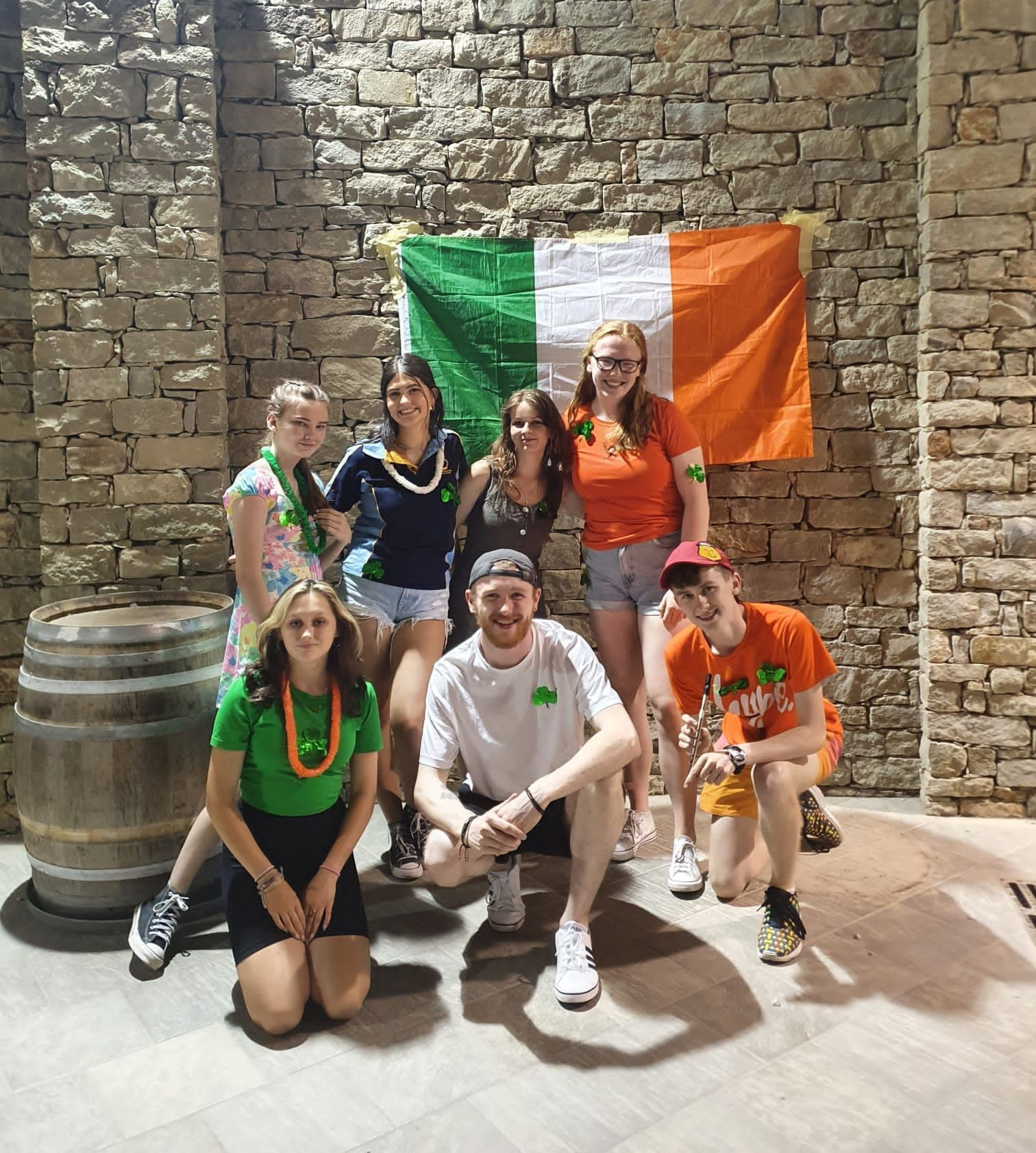 Grúpa ag seasamh laistigh os comhair bhratach na hÉireann, le héadaí féile glasa, bána agus oráiste.