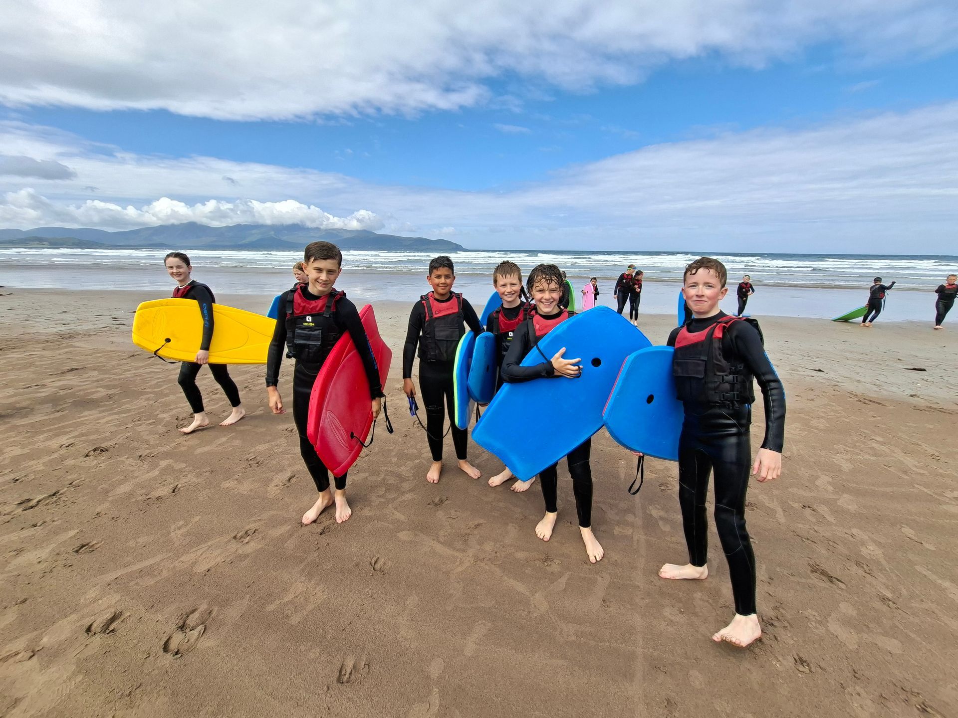 Surfálaithe ag iompar boird ildaite ar thrá ghainmheach faoi spéir scamallach