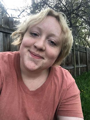 A woman in a pink shirt is smiling in front of a wooden fence.