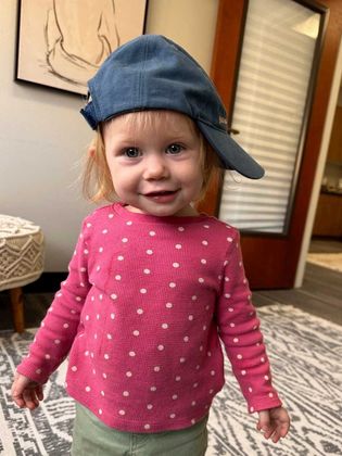 A little girl wearing a hat and a pink shirt is standing on a rug.