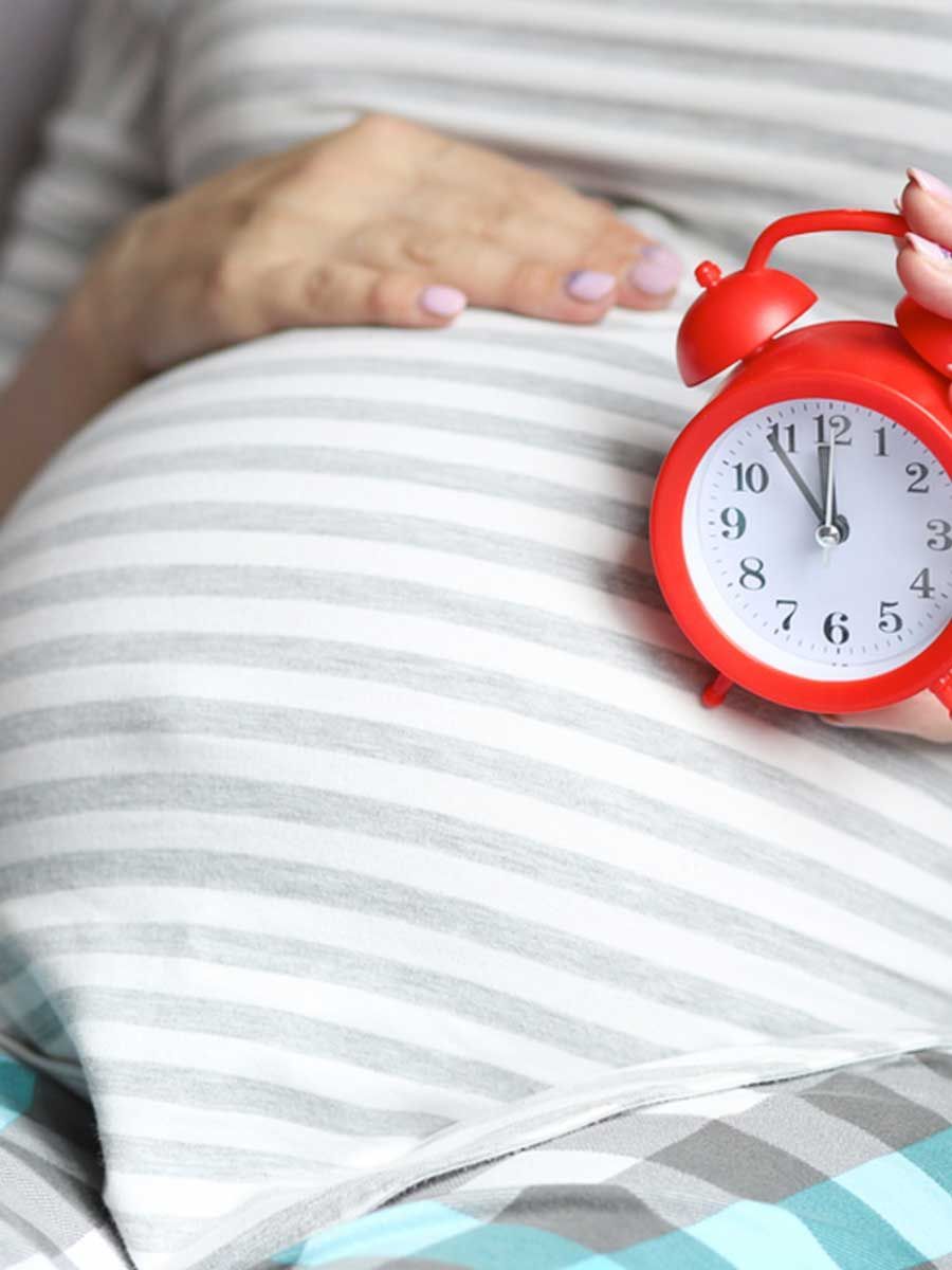 A pregnant woman is holding a red alarm clock in front of her belly.